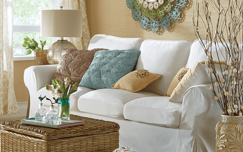A white sofa with teal and beige accent pillows, a wicker storage table, a beige floor vase with twigs, and vased flowers.