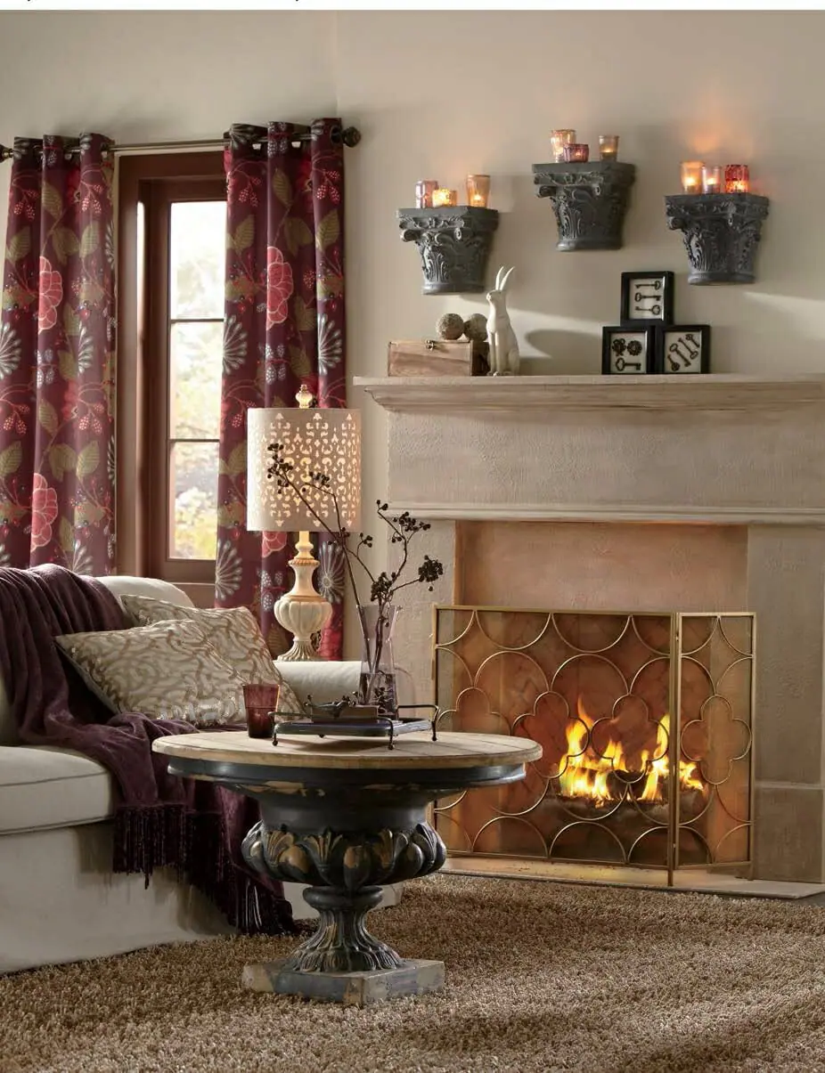 Lit votive candles on shelves above a fireplace, a pedestal coffee table, and a burgundy throw on an ivory sofa.