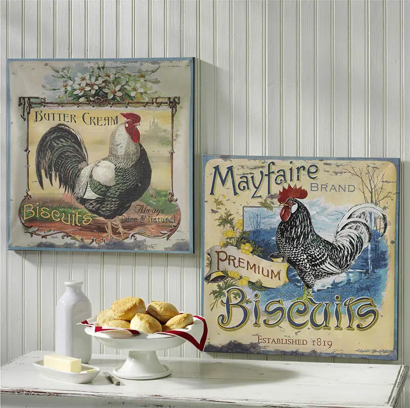 A set of two vintage rooster wall canvases on a white wainscot wall, by a cabinet topped with homemade biscuits.