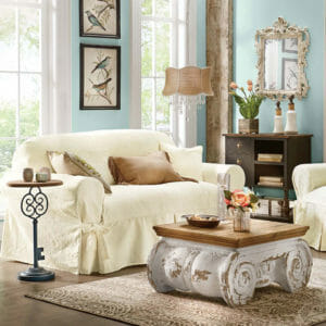 Ivory slipcovered sofa and chair with a scroll faux cement coffee table with vased flowers, and a skeleton key side table.