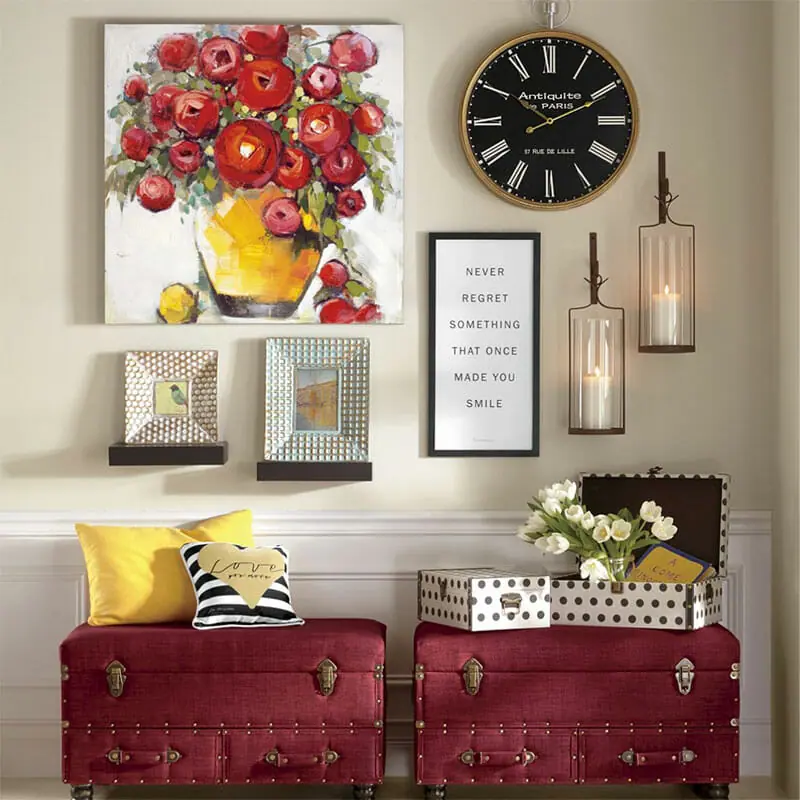 Two red trunk benches with pillows, a large canvas of red roses in a yellow pot, a round black clock, and two lit sconces.