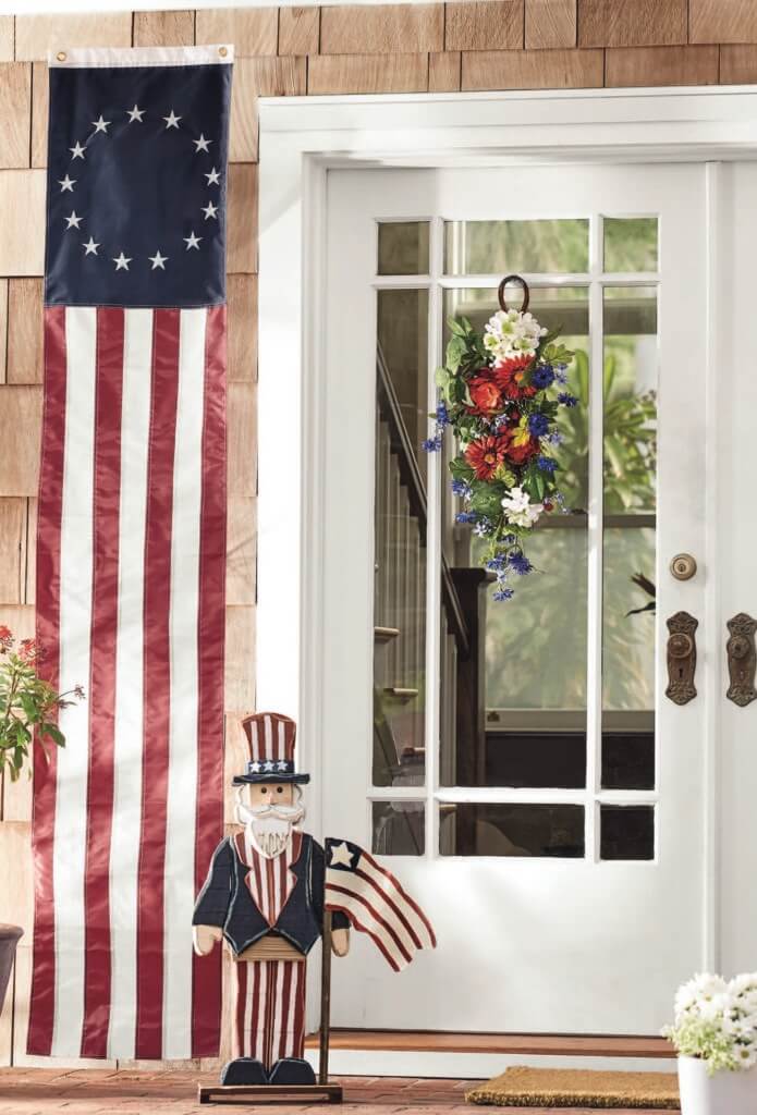 A patriotic porch with a red, white, and blue swag on a white door, a US 13-star banner, and a wooden Uncle Sam.