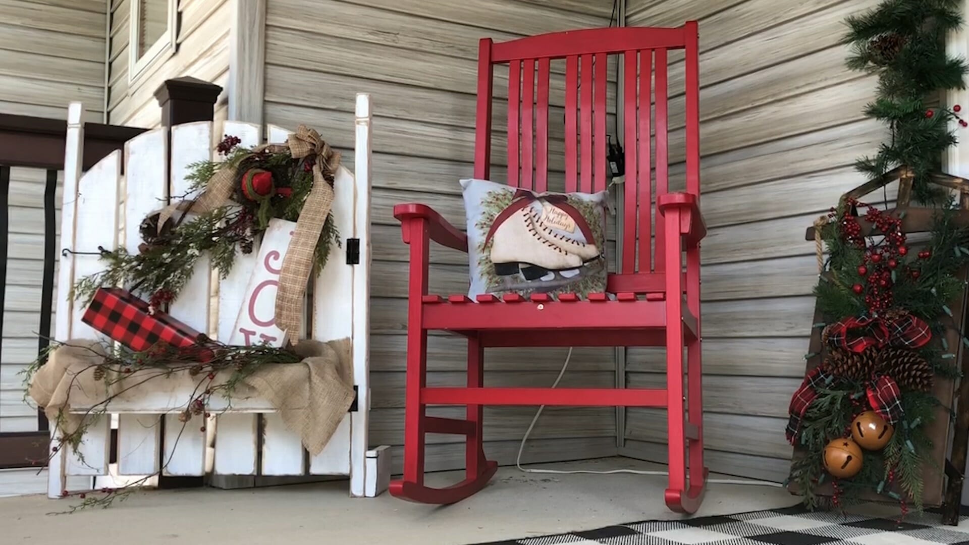 Porch with red rocking chair, ice skates pillow, a vintage sled and a white gate decorated with pine, ribbon, and bells.