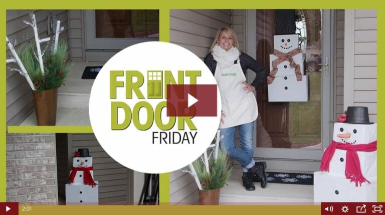 woman with a white scarf stands on a front porch adorned with a snowman wreath on front door and other winter décor