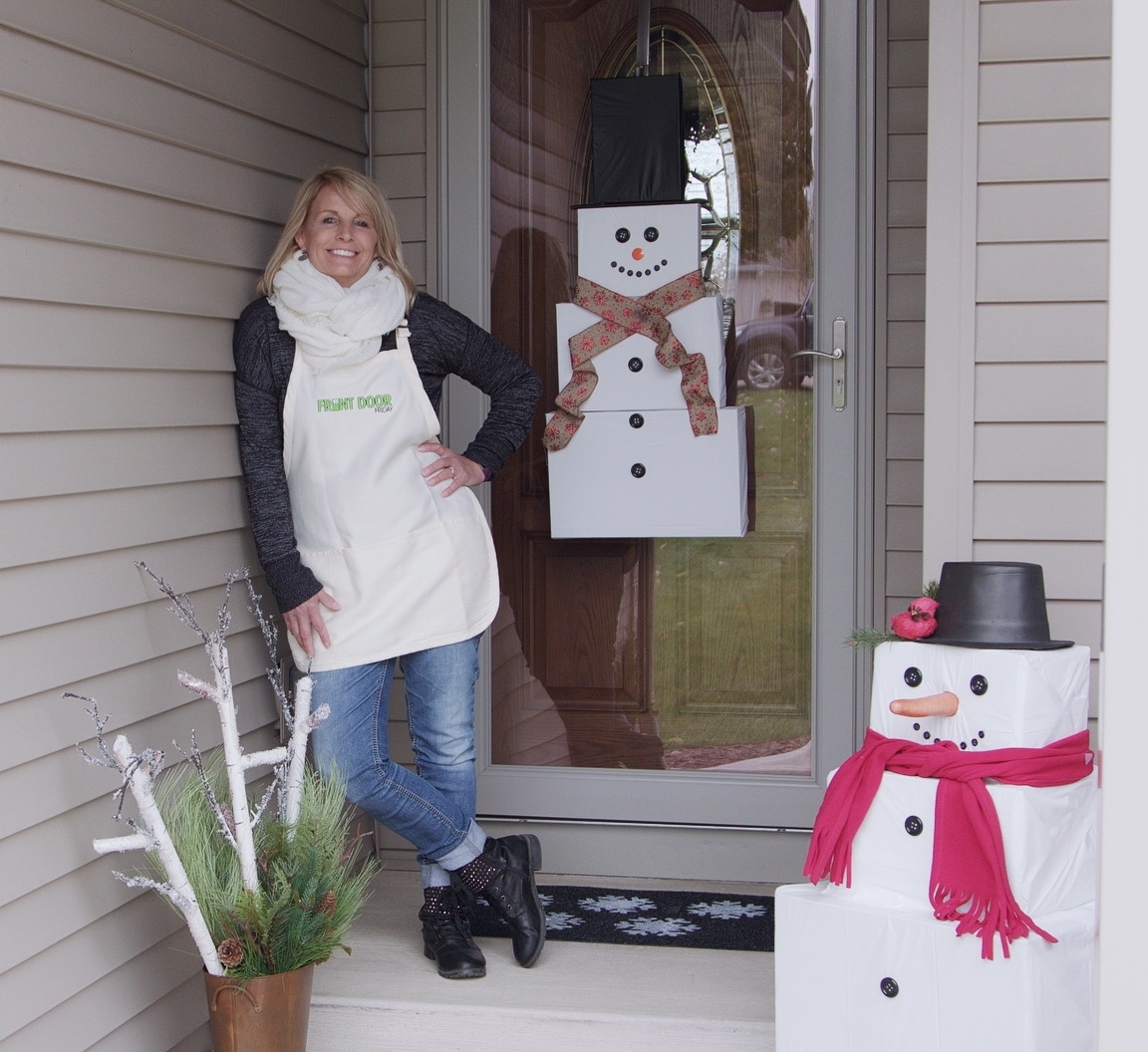 woman on porch with snowman hanging on door, sitting in corner and evergreens and birch branches in pot by steps