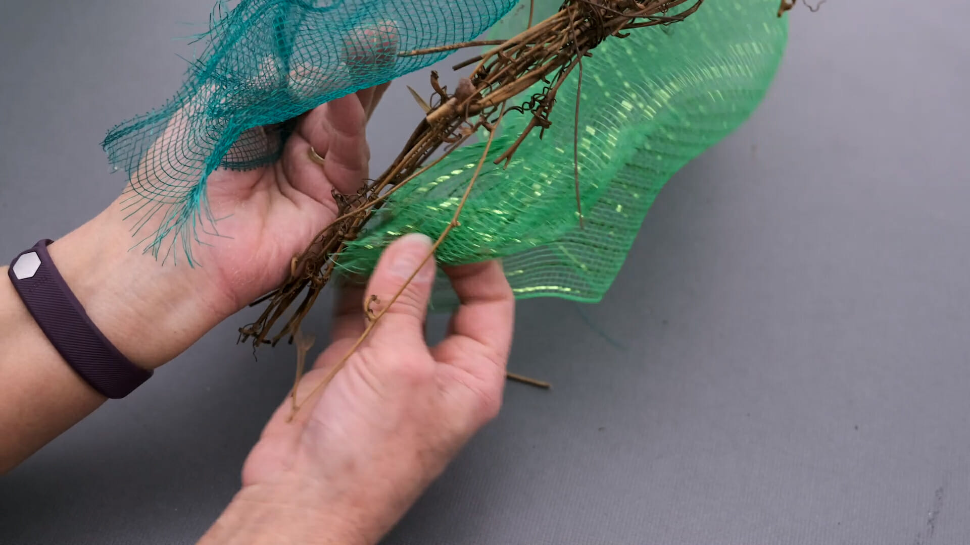 Hands wrapping two shades of green plastic netting onto twisted grapevine branches.