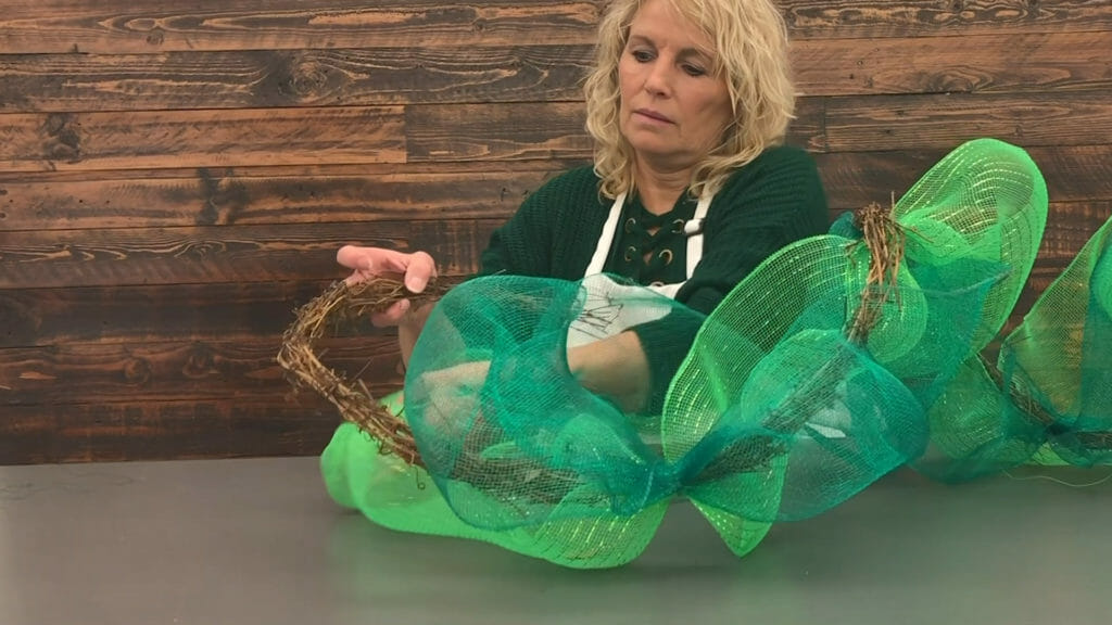 A blonde woman attaching two shades of green netting to a narrow grapevine garland.