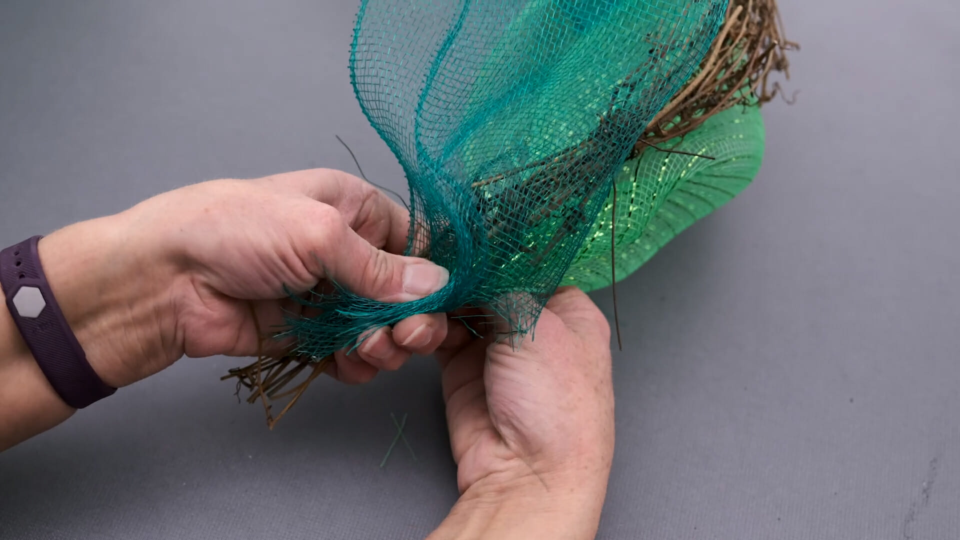 Close-up view of hands attaching two shades of green netting to a narrow grapevine garland.