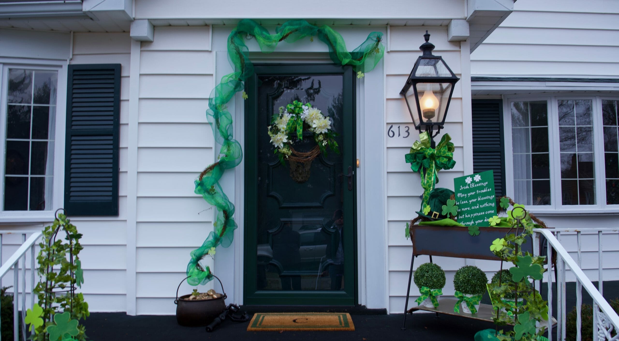 St. Patrick's Day front porch display of green garland landing in a pot of gold, a lit lantern with green bow, and planters.