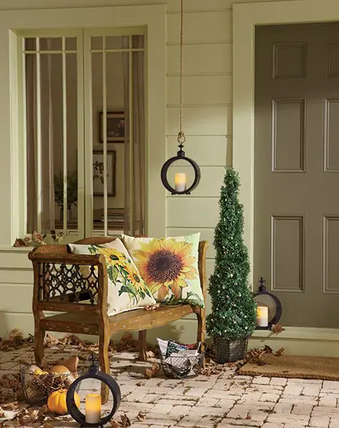 A wood porch bench with two sunflower pillows, and three black metal circle candle holders with lit candles.