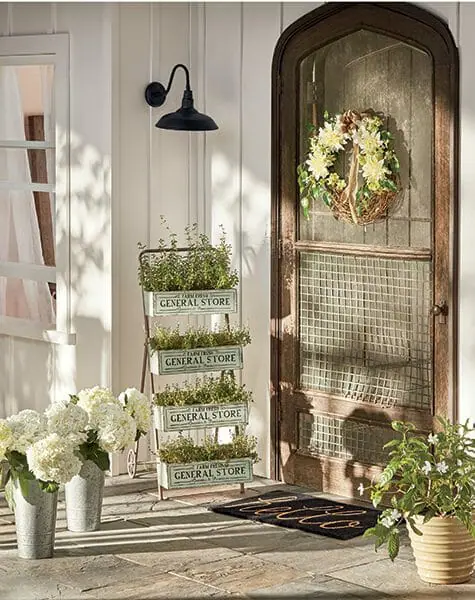 Farmhouse wood screen door with white floral wreath, tiered plant stand filled with herbs, and white hydrangeas in buckets.
