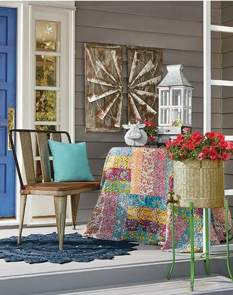 Farmhouse porch with quilt on table, rusty wash tub bench, windmill wall art, white lantern, metal bucket with red petunias.