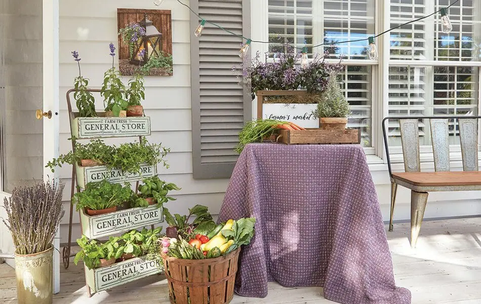 A Farmhouse porch with a tiered herb planter, a bushel basket of vegetables, a planter of purple flowers on a metal table.