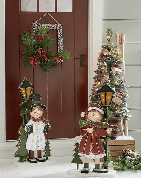 Two lit caroler cutouts in front of a red door hung with a square grapevine wreath with pine, red berries, and pine cones.