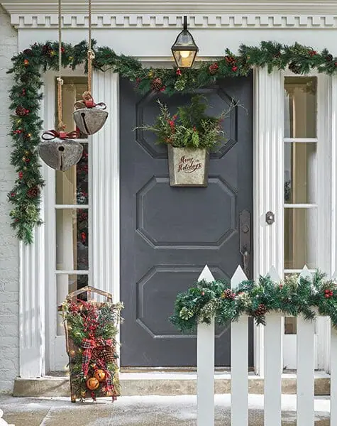 Gray front door with galvanized basket of greens, holiday garland, large hanging jingle bells, and a decorated sleigh.