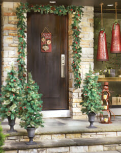 A front door hung with garland and Holiday plaque, three trees in urns, red bells, and a red metal snowman.