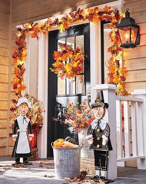 A porch with Autumn leaves garland and wreath, wooden Pilgrims, galvanized cans of gourds and dried florals