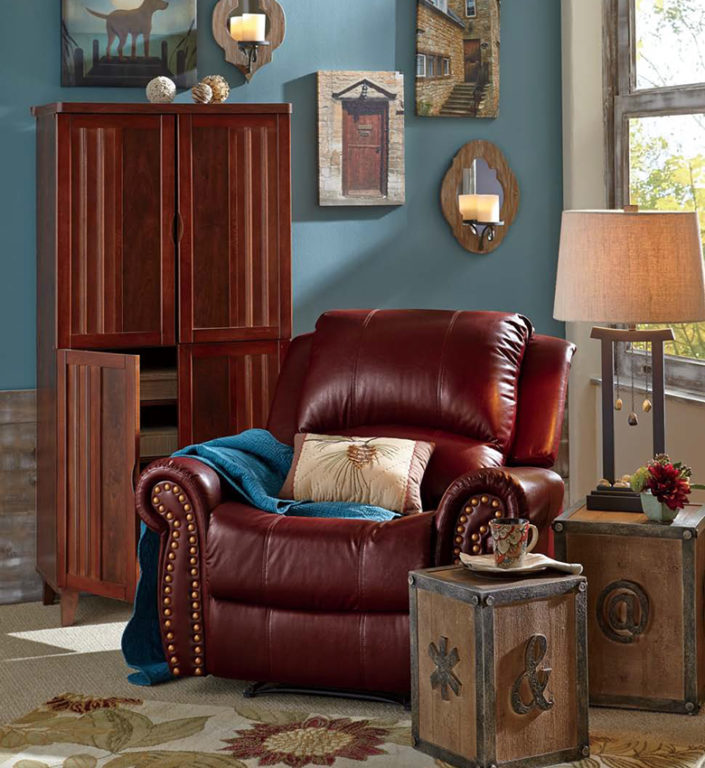 A brown nailhead recliner by a wood armoire, two rustic side tables, a sunflower area rug, and teal walls. 
