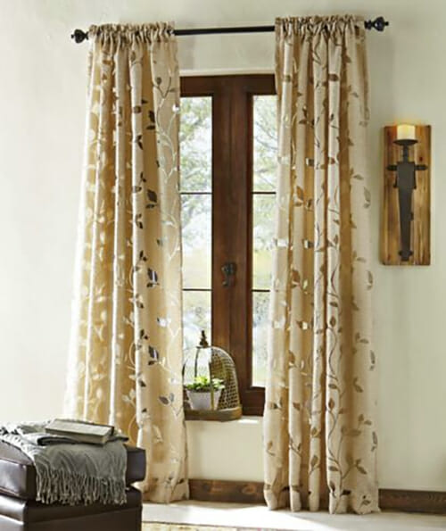 Long beige curtains with a silver vines print on a window with a plant on the ledge, near a lit sconce.