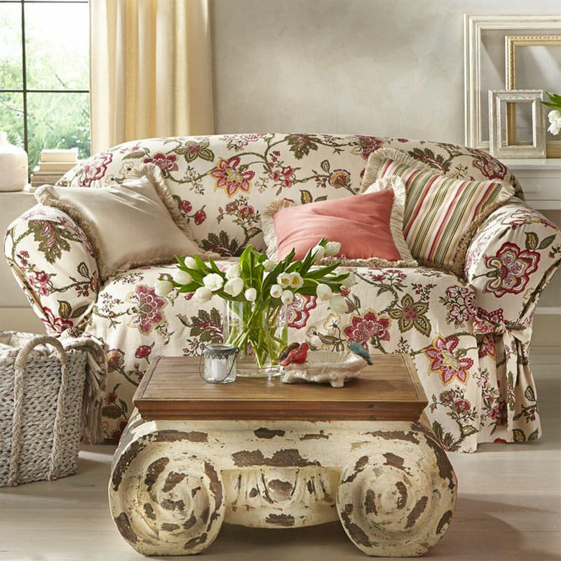 A loveseat in an ivory and rose floral slipcover, accent pillows, and a scrolled faux cement coffee table with white tulips.