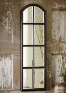An eight-paned wood framed mirror with a top arch placed on weathered paneling.