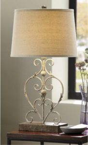 A scrolled weathered-look metal table lamp with a lit beige shade, next to a lavender glass vase of wildflowers.