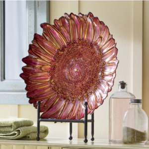 A 3D red and yellow glass bowl in the shape of a sunflower, placed on a black metal stand by two glass bottles.