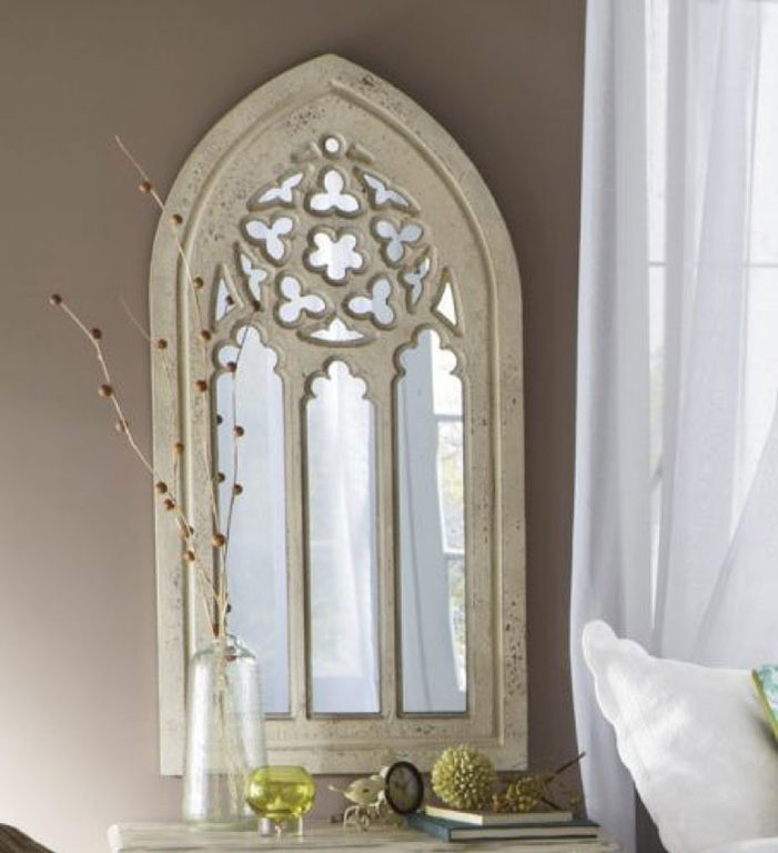 A rustic cathedral shaped wall mirror in ivory, with a cutout top medallion.
