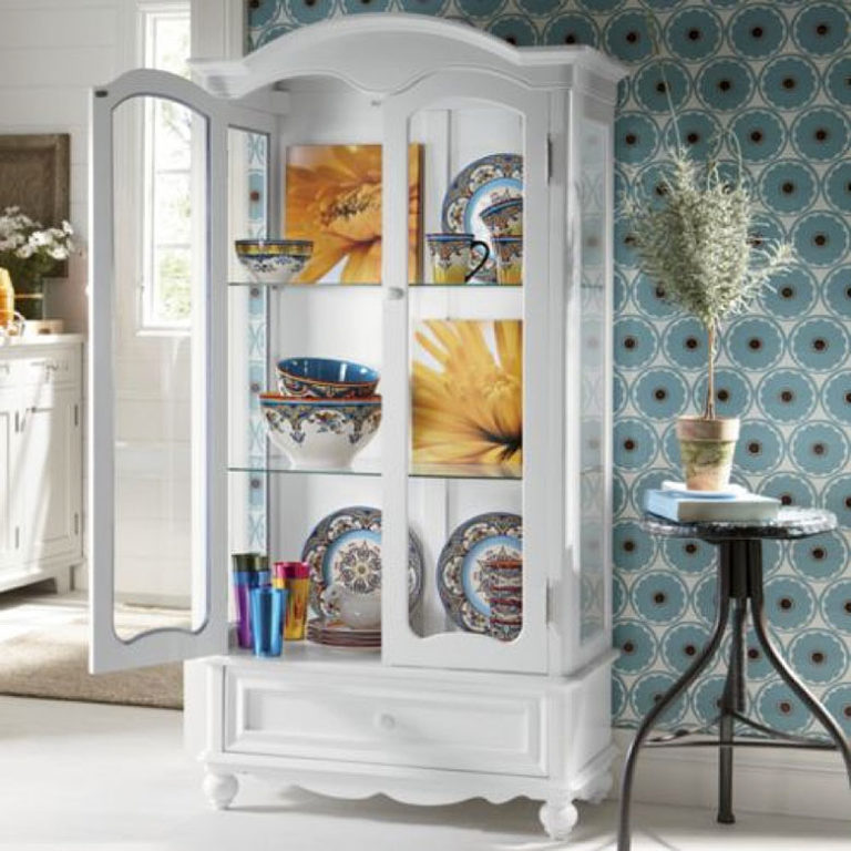 A tall white Victorian-look display cabinet with two glass doors and a bottom drawer, filled with floral dinnerware.