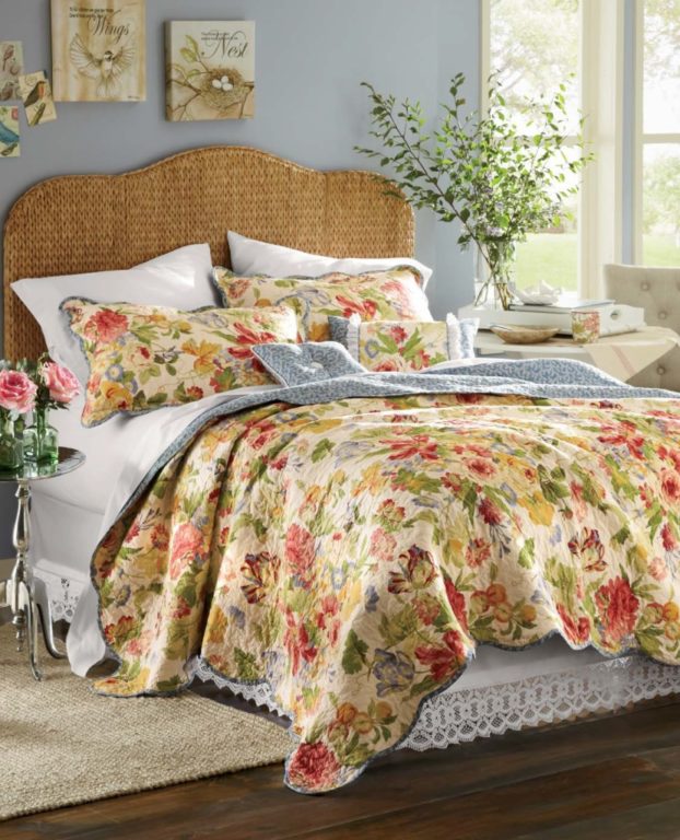 A multicolor floral quilt with reversible blue and white back, white sheets, a seagrass headboard, and bird canvases.