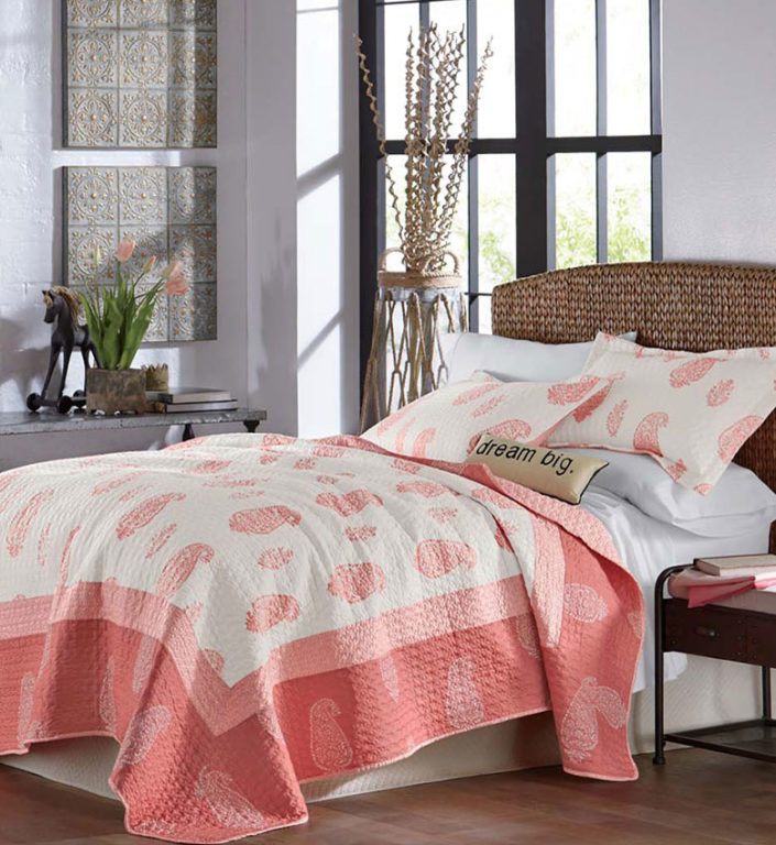 A white and coral paisley quilt with a seagrass headboard, embossed gray plaques on a white wall, and coral tulips.