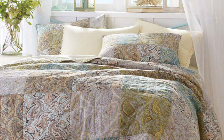 A quilt with pastel paisley squares over a beige sheet set, vased white flowers, lanterns and coral by open windows.