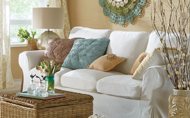 A white sofa with teal and beige accent pillows, a wicker storage table, a beige floor vase with twigs, and vased flowers.