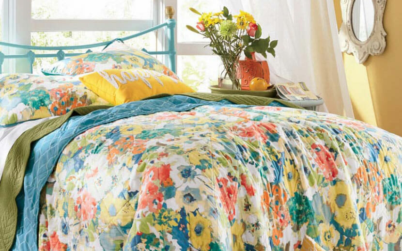 A Spring multicolor floral bed set in teal, yellow, and orange, next to a side table with vased flowers by an open window.