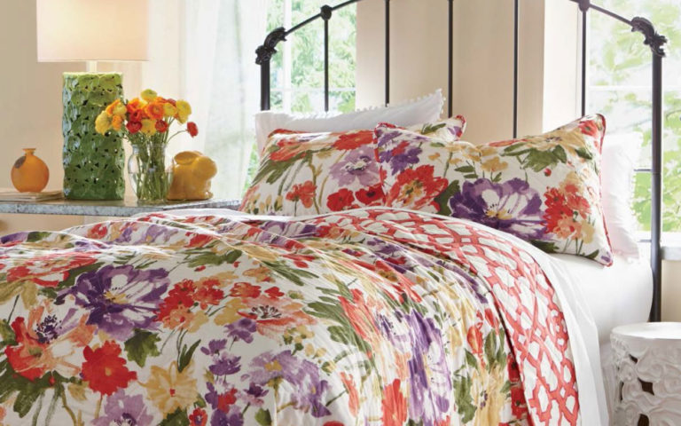 A black iron bed topped with a multicolor floral bedspread and shams, by a lit table lamp and vased flowers.
