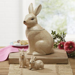 An ivory sitting rabbit cookie jar on a table with two bunny salt and pepper shakers, with a plate of cookies.