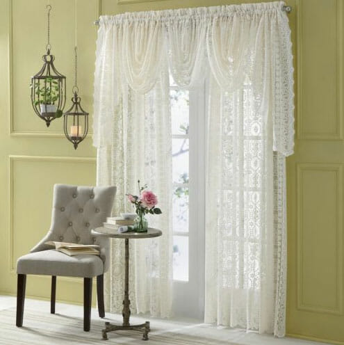 White lace curtains and valances, a beige tufted chair, a side table with vased roses and two hanging lanterns.