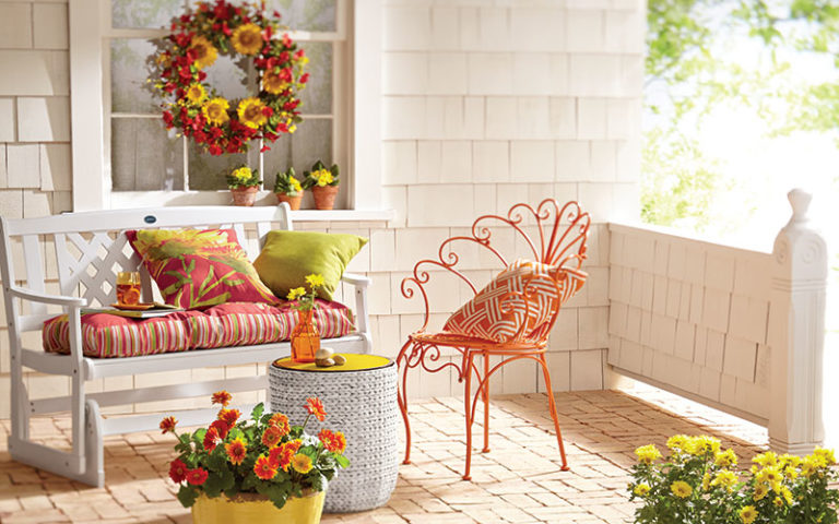 white bench with green and orange cushions next to orange metal chair on porch with orange and yellow potted flowers