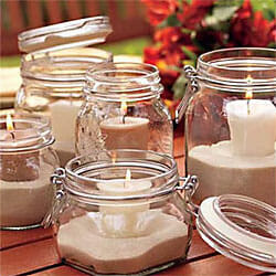 Five different clear glass jars with lit white candles placed in sand on the bottom of each one.