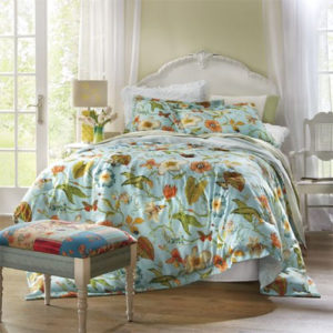 White headboard with a comforter set in a blue botanical print with butterflies, white sheer curtains, and quilt-top stool.