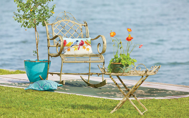A vintage white metal Bordeaux rocker with a colorful bird pillow, lakeside, by a young tree.