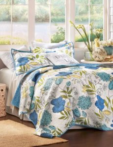 A white and blue floral quilt with matching shams and white sheets, against sunny windows and a table with flowers.