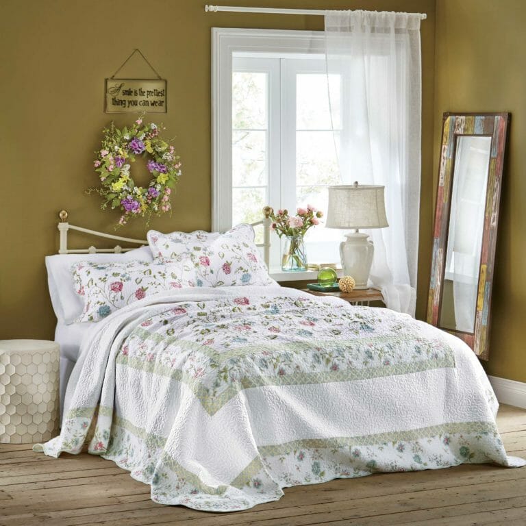 Bed with white and floral print shams and bedspread, white sheer curtain, round floral wreath, and tall rustic mirror.