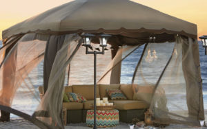 A tan gazebo on the beach with netting, lit lanterns, and a tan couch with multicolor zigzag pattern ottoman and pillows.