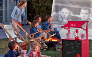 A family with four children outside by a firepit and sling chairs, watching an old movie on a sheet while eating popcorn.
