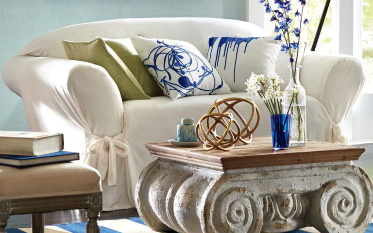 White slipcovered loveseat, white and blue swirl pillows, scroll faux cement coffee table, and vased blue and white flowers.