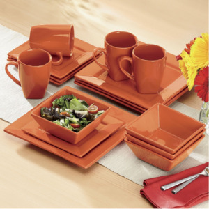 Square rust-colored dinnerware set on a beige table runner, next to vased flowers.