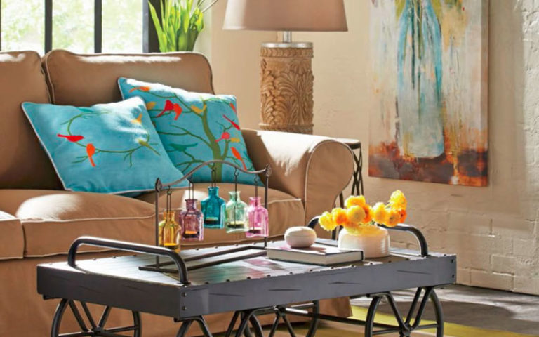 A tan sofa with two blue pillows with birds, a tall canvas of flowers in a blue vase, a beige lamp, and a cart coffee table.