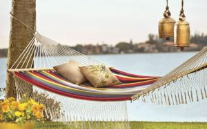 A multicolor stripe hammock with fringe tied to a tree next to a lake, with two hanging brass bells, and yellow potted mums.