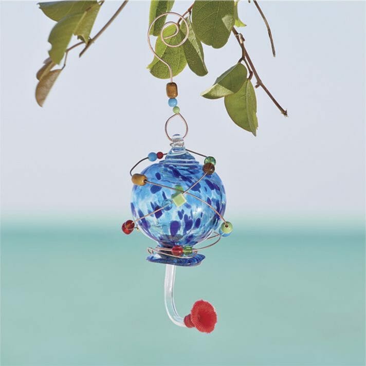 A blue glass hummingbird feeder wrapped with multicolored beads, hanging from a branch by coiled copper wire.